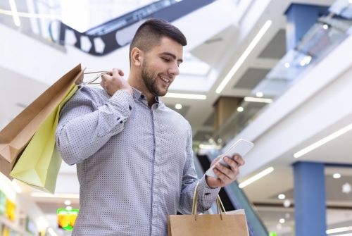 Happy guy with cellphone and purchases in shopping center